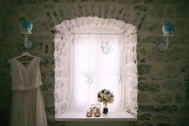El vestido de novia por la ventana con el ramo de novias y sandalias en el alféizar. - Foto, imagen