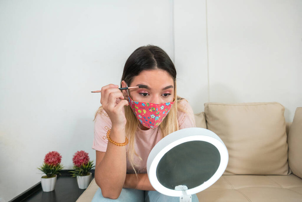 Женщина в коронавирусной защитной маске и макияже. Женщина в маске смотрит на себя в зеркало и наносит макияж. Медицинская маска, крупный план, профилактика COVID-19 - Фото, изображение