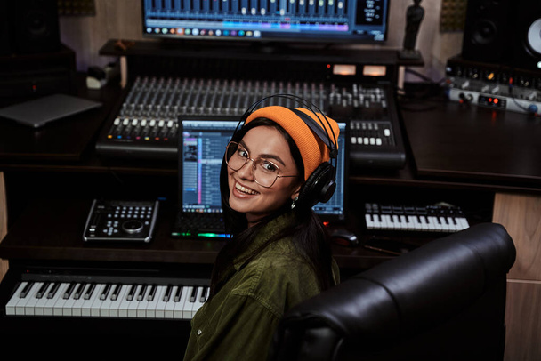 Portrait de belle jeune brune, artiste féminine souriante à la caméra tout en jouant synthétiseur clavier, assise en studio d'enregistrement - Photo, image