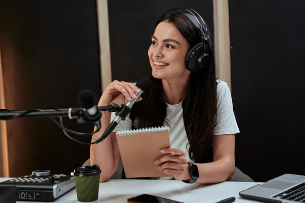 Çekici genç bayan radyo sunucusunun portresi mikrofonla konuşurken bir yana gülümsüyor, canlı yayını düzenliyor. - Fotoğraf, Görsel