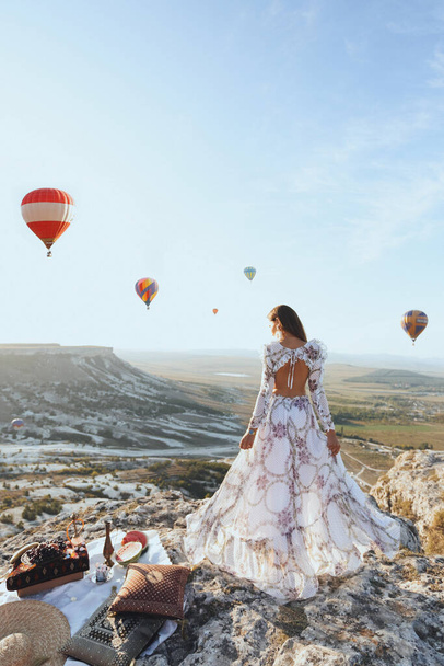 fashion outdoor photo of beautiful woman with dark hair in elegant dress having picnic with fantastic view on valley with air balloons on background - Foto, Imagem