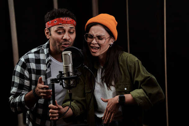 Fiatal férfi és nő portréja, duett énekel egy kondenzátor mikrofonba, miközben egy dalt rögzít egy professzionális stúdióban - Fotó, kép