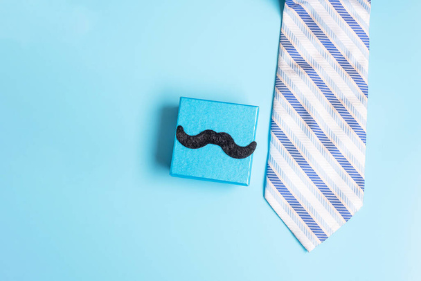 Geschenkbox mit Schnurrbart und Krawatte auf blauem Hintergrund, Vorbereitung für Vater. Weltmännertag und Vatertagskonzept - Foto, Bild