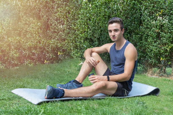 joven atleta de fitness sentado en la alfombra y descansando tomando un pequeño café y un bar de energía para relajarse y recuperarse en un día soleado - Foto, imagen