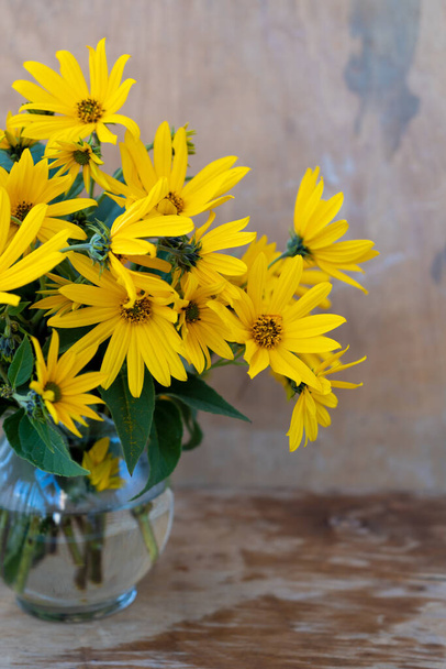 A bouquet of yellow Jerusalem artichoke flowers in a glass vase on a wooden background. Postcard with place for text. A simple wild flower that looks like a chamomile. - Foto, imagen