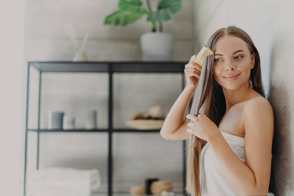 Attractive young woman smiles and enjoys her beauty, combes long straight hair after applying mask, stands wrapped in bath towel in cozy bathroom, has smooth skin. Hair care, spa and beauty concept - Photo, Image