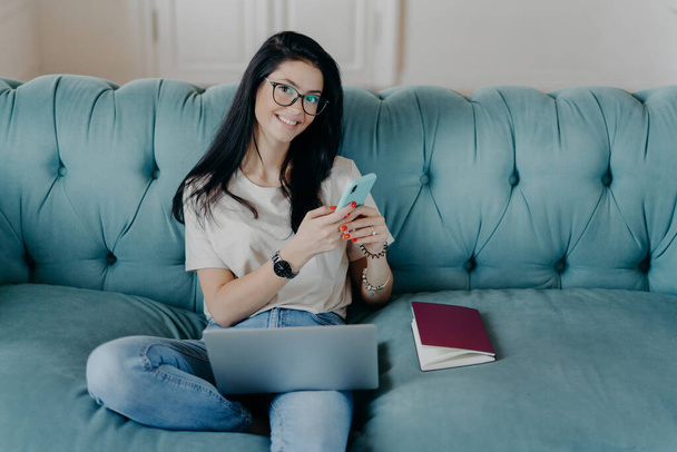 Happy relaxed European woman student studies distantly, uses modern gadgets, creats new project or report, dressed in t shirt and jeans, poses on comfortable couch, updates software. View from above. - Photo, Image
