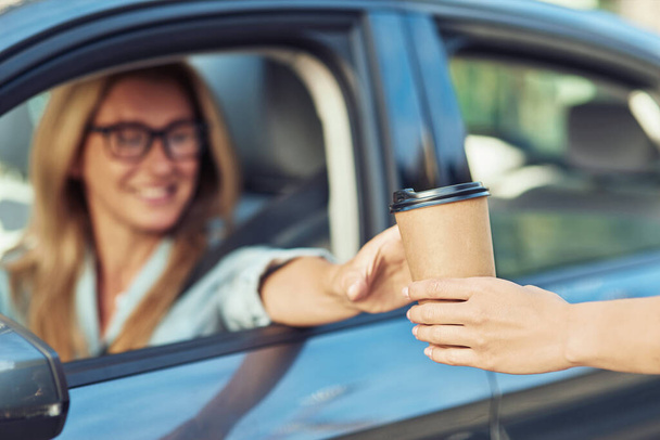 Ama önce kahve. Kafkasyalı mutlu iş kadını modern arabasının direksiyonunda oturuyor ve kahve ısmarlıyor, kağıt bardağa odaklanıyor. - Fotoğraf, Görsel