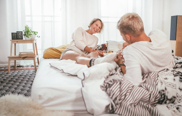Laughing blonde hair young adults couple in pajamas lazy relaxing lying in a cozy bed in the bedroom and having a peacefully carefree chatting. Couples relations concept image. - Foto, imagen