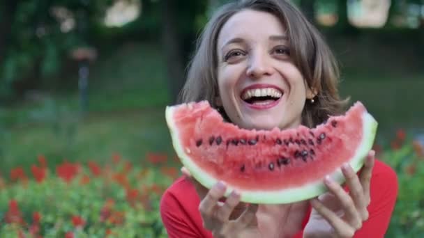 Schöne Frau in rotem Hemd mit kurzen Haaren sitzt draußen auf rotem Blumenhintergrund und isst ein Stück Wassermelone. Picknick in der Stadt. - Filmmaterial, Video