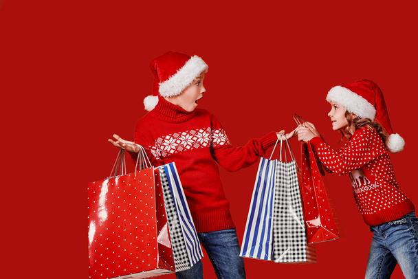 Cute little girl in Santa hat hanging shopping bag with Christmas gifts on hand of astonished boy during holiday celebration against red background - Foto, afbeelding
