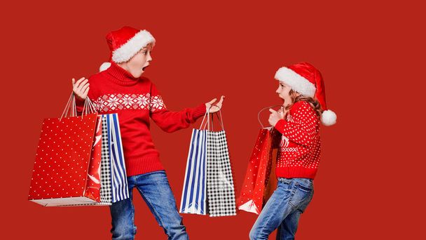 Cute little girl in Santa hat hanging shopping bag with Christmas gifts of astonished boy during holiday celebration against red background - Photo, image