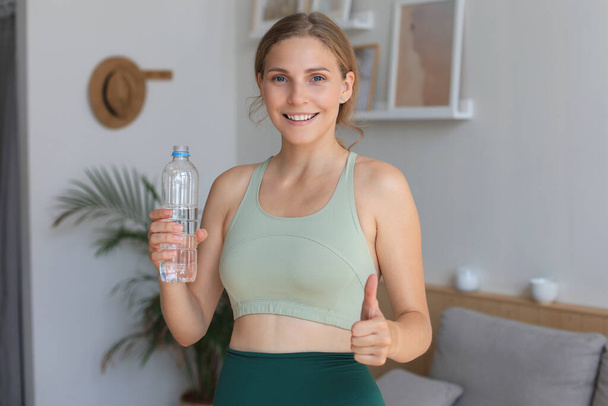 Young beautiful woman standing with dumbbell and drink water after exercising looking camera. Attractive female bodybuilder working out. Fitness and healthy lifestyle concept - Foto, Imagem