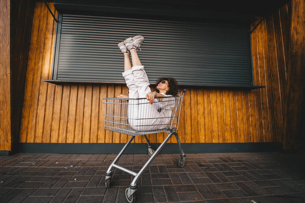 Souriant jeune fille brune dans des lunettes de soleil avec les jambes levées assis à l'intérieur d'un chariot d'achat. Portrait horizontal - Photo, image