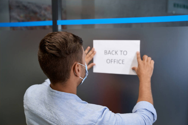 Visão traseira de um jovem, trabalhador de escritório usando máscara protetora colando sinal aberto com texto de volta ao escritório na porta de vidro - Foto, Imagem