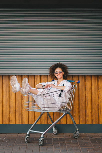 Μόδα δροσερό χαμογελαστό κορίτσι με μαλλιά afro και γυαλιά ηλίου διασκεδάζοντας κάθεται στο καροτσάκι ψώνια. Κάθετη απεικόνιση - Φωτογραφία, εικόνα