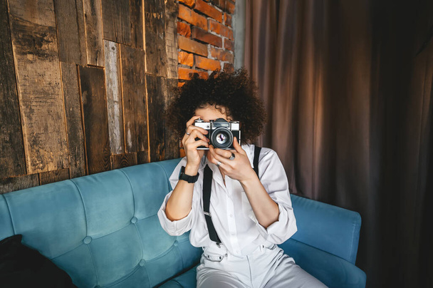 Jolie jeune femme souriante aux cheveux afro portant une chemise assise sur un canapé bleu au studio, tenant un appareil photo. - Photo, image
