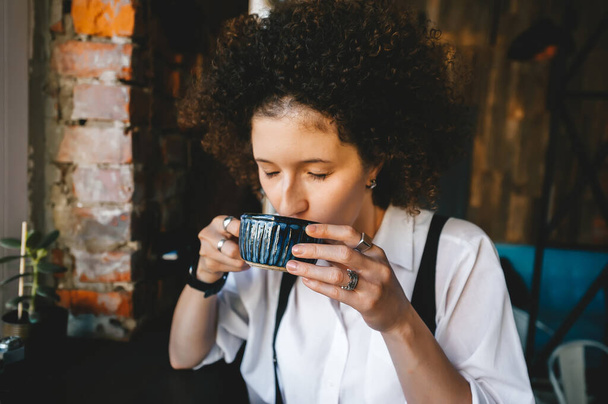 Μελαχρινή γυναίκα με αφρο μαλλιά και με κλειστά μάτια κάθεται στο καφέ με ζεστό καφέ. Κλείσιμο. - Φωτογραφία, εικόνα