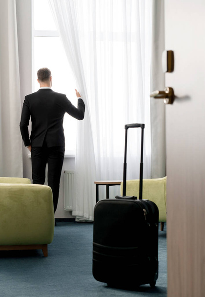 Вид сзади на бизнесмена в темном костюме, стоящего в гостиничном номере и смотрящего в окно - Фото, изображение