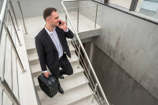 Νεαρός επιχειρηματίας με βαλίτσα στο κομψό κοστούμι μιλώντας στο τηλέφωνο, ενώ περπατάτε από τις σκάλες στο σύγχρονο κτίριο των επιχειρήσεων - Φωτογραφία, εικόνα