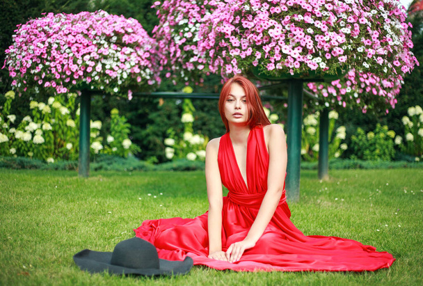 Young attractive girl in a long red dress with a black hat, in a flower garden on the lawn - 写真・画像