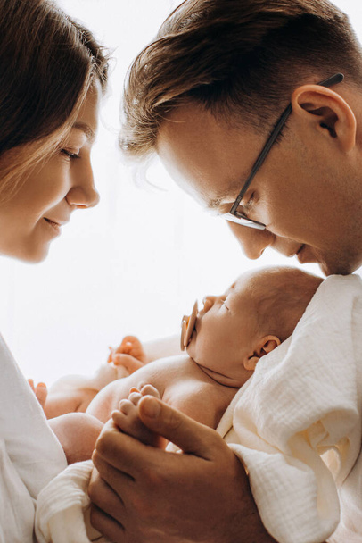 Beautiful parents with newborn baby, loving mom hold little daughter in arms, caring dad gently hold a hand of adorable baby girl, enjoy tender family moments, parenting concept - Фото, изображение