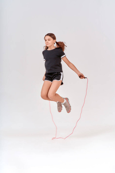 Здоровый и активный. Полнометражный снимок милой девочки-подростка, прыгающей с скакалкой на сером фоне в студии - Фото, изображение
