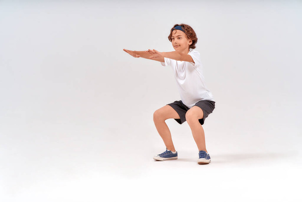 physical activities. Full length of caucasian teenage boy in sportswear squatting, doing sit-ups isolated over grey background in studio - Foto, imagen