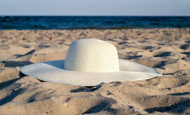 Yumuşak kumun üzerinde denizin arka planına karşı duran büyük beyaz hasır şapka. Plaj aksesuarları. Seyahat ve tatil kavramı. - Fotoğraf, Görsel