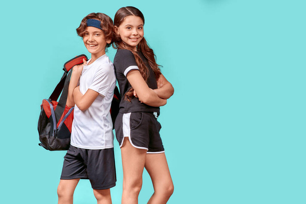 Готовимся к совместной тренировке. Два счастливых подростка мальчик и девочка в спортивной одежде смотрят в камеру и улыбаются, стоя спиной к спине изолированы на синем фоне - Фото, изображение