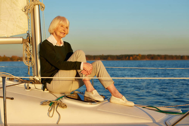 Ηρέμησε και χαλάρωσε. Όμορφη ηλικιωμένη γυναίκα που κάθεται στην πλευρά του ιστιοφόρου ή κατάστρωμα γιοτ επιπλέουν στην ήρεμη γαλάζια θάλασσα στο ηλιοβασίλεμα, απολαμβάνοντας καταπληκτική θέα και χαμογελώντας - Φωτογραφία, εικόνα