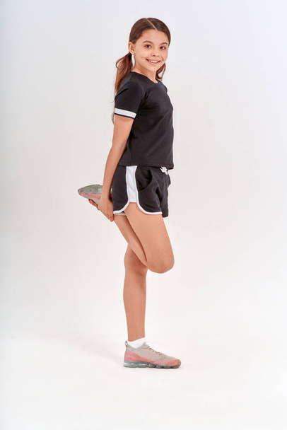 Разминка перед тренировкой. Полная длина симпатичной девочки-подростка в спортивной одежде, растягивающей ногу, находясь в изоляции на сером фоне в студии - Фото, изображение