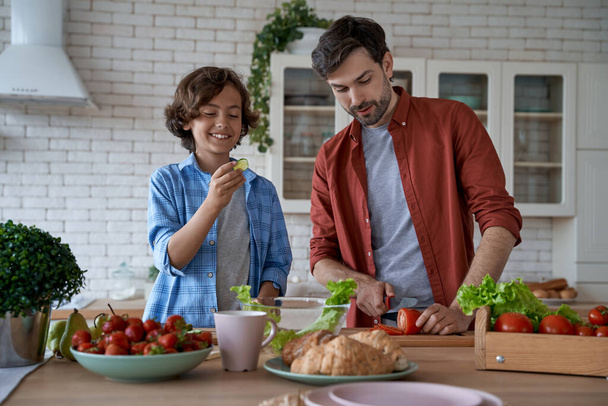 Das Wochenende zu Hause genießen. Junger Vater und kleiner glücklicher Sohn schneiden frisches Gemüse und bereiten gemeinsam in der modernen Küche einen Salat zu - Foto, Bild