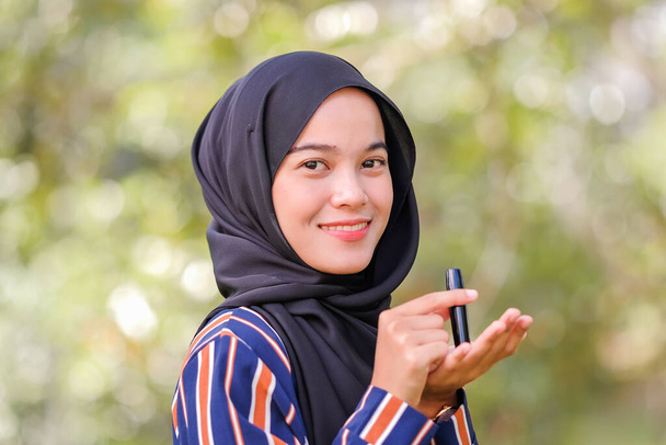 Χαμογελαστή όμορφη νεαρή μουσουλμάνα γυναίκα φορώντας μαντίλα και κρατώντας ένα κραγιόν σε θολή φόντο. - Φωτογραφία, εικόνα