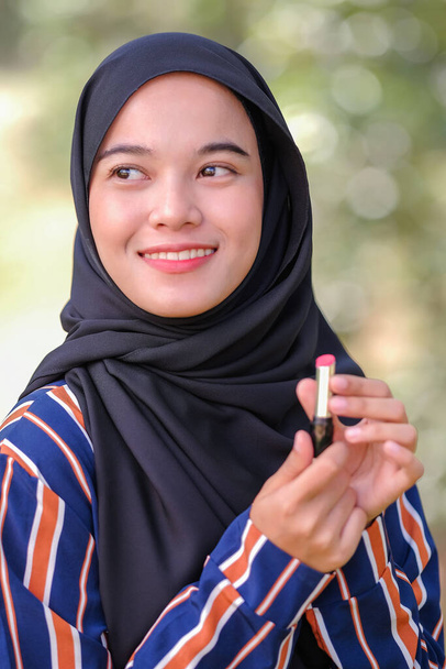 Smiley mooie jonge moslim vrouw dragen hijab en het houden van een lippenstift op wazig achtergrond. - Foto, afbeelding