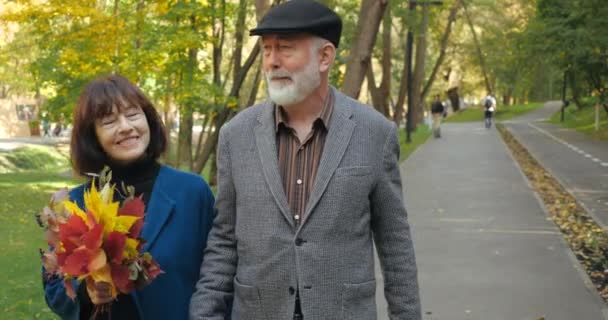 Aantrekkelijk glimlachend bejaard echtpaar met baard man en vrouw met een boeket herfstbladeren genieten van een actieve wandeling in het park. Vrolijke grootouders lopen buiten in de frisse lucht, in de buurt van front view. - Video