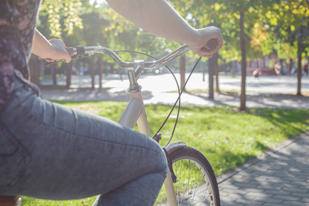 A girl in jeans and sneakers rides a beige bike in the park among green trees and lawn - 写真・画像