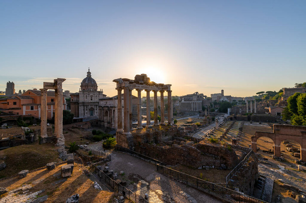 Cichy świt w Forum Rzymskim, Rzym. Promienie słoneczne pojawiają się w świątyni Saturna i oświetlają budynki, łuk Severusa, świątynię Westy, łuk Tytusa, Koloseum. Włochy. - Zdjęcie, obraz