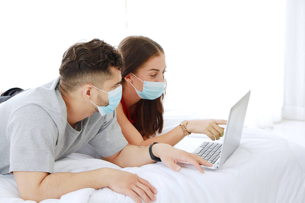 Beyaz çift Covid 19 virüsüne karşı maske takmayı ve sosyal medyada dizüstü bilgisayarla alışveriş yapmayı ve beyaz yatakta yan yana yatmayı seviyor. Tatil konseptinde teknoloji ve yaşam tarzı. - Fotoğraf, Görsel
