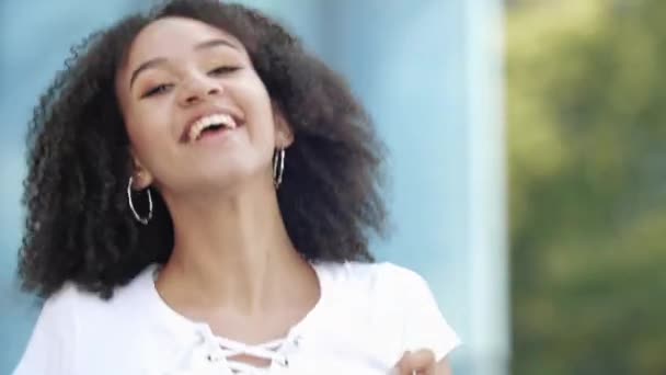 Cheerful beautiful cute curly afro american young woman enjoys jumping holding smartphone, waving arms from exciting emotions, laughing joyfully and smiling with happiness in modern urban city street. - Footage, Video
