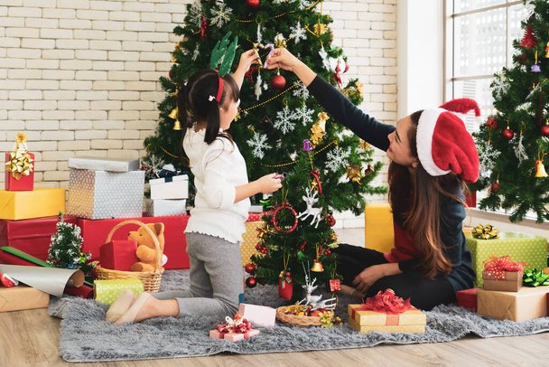 asian mother and her young daughter wearing santa claus hat and using ornament to decorate christmas tree. family together concept - Фото, зображення