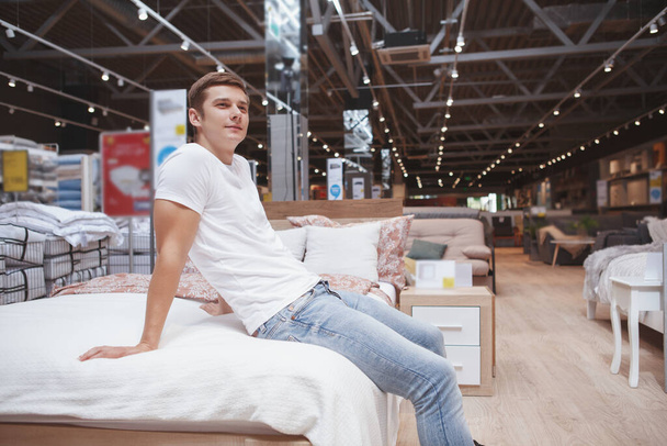 Handsome young man sitting on a new be on sale at furniture supermarket - Foto, Bild