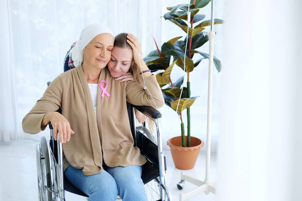 Caucasian young daughter Hug With love and care for her mother in white headscarf is sitting on wheelchair after chemotherapy. Elderly mother is suffering from cancer or Leukemia patient. - Valokuva, kuva