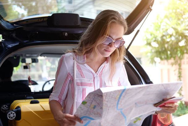 Hymyilevä nuori valkoihoinen matkustaja nainen on yllään aurinkolasi ja istuu auton vuokrattavana. Hänellä on kartta löytää matkustaa kaupungin ulkomaisen pääoman Bangkokissa, Thaimaassa. Maantiematkan käsite. - Valokuva, kuva