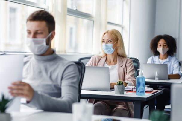 Teamwork on project. Multiracial employees in protective masks work on laptops at desks with antiseptics, focus on lady, looking out the window - Foto, Bild