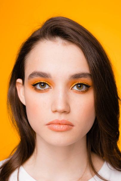 Portrait of a girl of European appearance with bright eye makeup, stands on an orange background - Фото, изображение