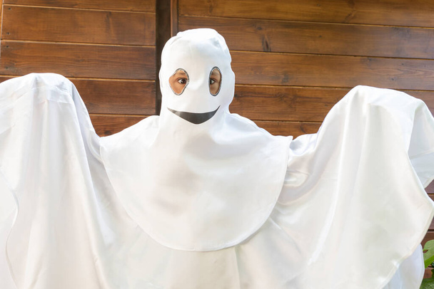 garçon en costume de fantôme blanc avec sourire est prêt pour la fête d'Halloween sur fond en bois. Concept d'Halloween. Espace de copie pour le texte. - Photo, image
