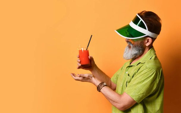 Bearded man in green sun visor, shirt and sunglasses. Holding glass of squeezed juice, clenched fist, posing on orange background - Valokuva, kuva