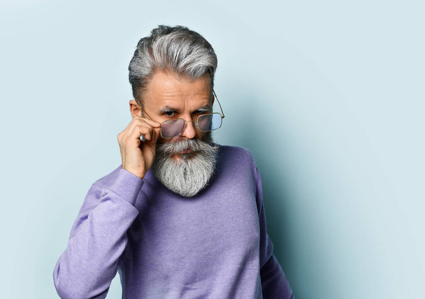 Gray-haired aged man in purple sweater. Fixing his sunglasses and looking at you while posing against blue background. Close up - Foto, Imagem