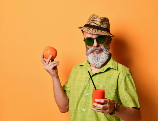 Aged man in hat, green shirt, sunglasses. Smiling, holding an orange, glass of fresh juice with tube, posing on orange background - Zdjęcie, obraz